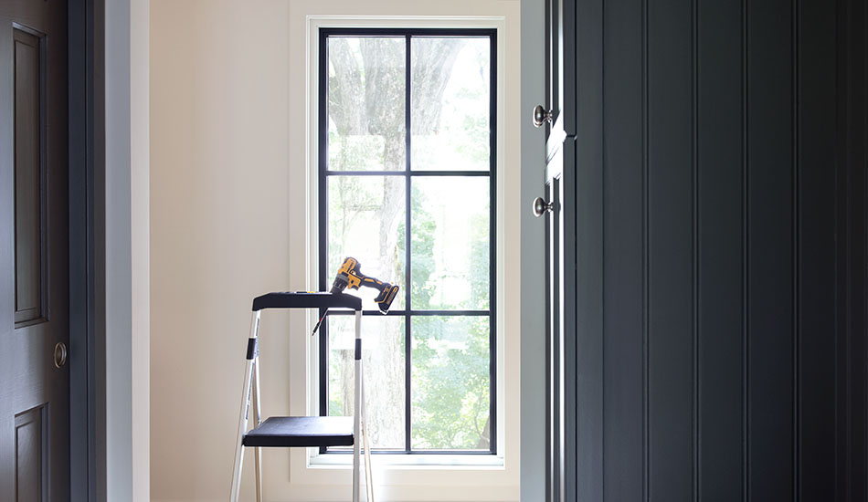 A stepladder with a drill resting in a holder sits in front of a tall narrow window with a black frame