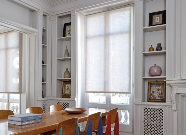 Tall windows featuring roller shades in a Dining Room with built in shelves and a wooden table & chairs with colorful accents