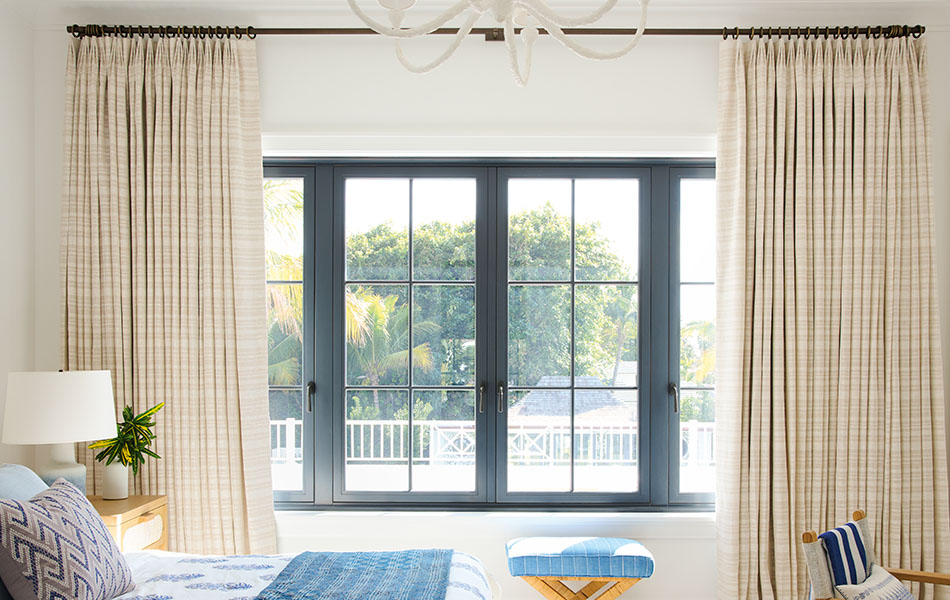 Beige-sand-colored thermal drapes are pulled apart to reveal trees and a pool while inside light falls on a crisply made bed