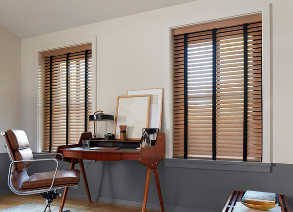 Two windows featuring wood blinds in an office with a brown chair and centered wood desk against grey and white walls